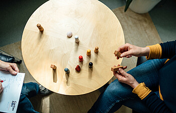 Von oben sieht man einem runden Tisch auf dem Spielfiguren stehen, eine Hilfesuchende hat zwei dieser Figuren in der Hand und die Beraterin sitzt mit einem Block auf der gegenüberliegenden Seite des Tisches.